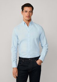 Camicia button-up di un delicato blu, realizzata in tessuto liscio, con colletto classico e logo sul petto, abbinata a jeans scuri.