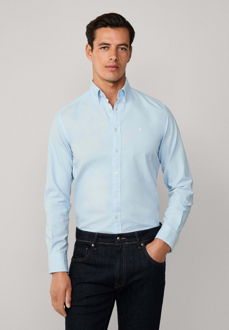 Camicia button-up di un delicato blu, realizzata in tessuto liscio, con colletto classico e logo sul petto, abbinata a jeans scuri.