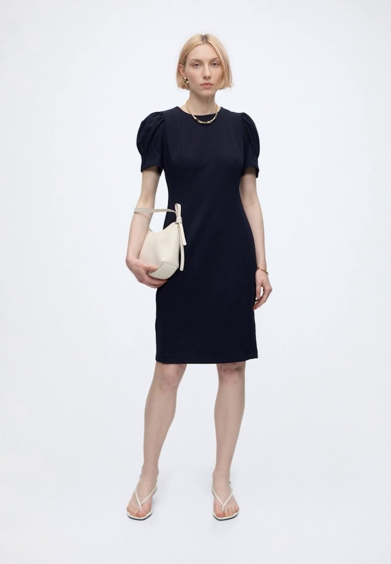 Femme debout portant une robe noire ajustée avec des manches bouffantes, tenant un petit sac à main blanc, et portant des sandales blanches sur un fond blanc uni.
