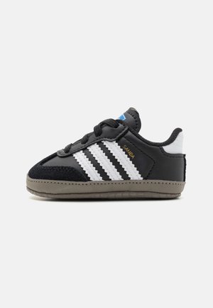 Czarnobiały but sportowy Adidas Samba z trzema białymi paskami, złotym napisem "SAMBA", czarnymi sznurówkami i szarą podeszwą na neutralnym tle.