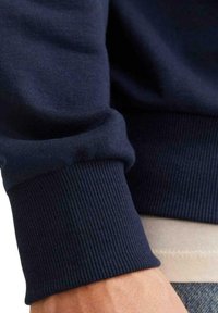 Puño de sudadera azul marino con textura acanalada, que presenta un diseño ajustado y suave. El puño se extiende sobre una camisa de color claro que lleva debajo.