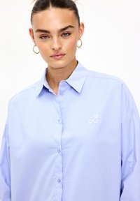 Camisa de collar azul claro con cierre de botones, corte holgado y sutil logo bordado de "L" en el pecho. Textura de tela suave.