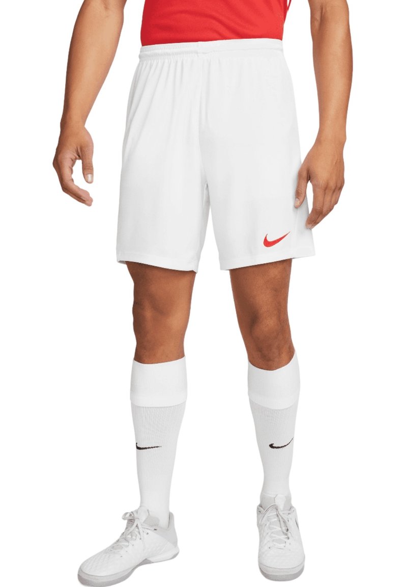 Homme portant un short de sport blanc Nike, des chaussettes blanches Nike, des chaussures de sport blanches et un t-shirt rouge, debout les bras détendus le long du corps.