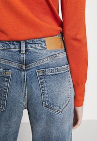 Blue denim jeans with a distressed texture and brown leather patch at the waistband, worn with an orange sweater, showing the back view.