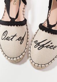 Espadrilles en toile avec coutures et bordures noires, avec le texte manuscrit noir « Out of office » sur les orteils.