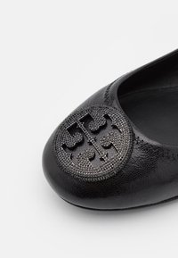 Chaussure plate en cuir noir avec emblème rond en métal texturé du logo sur l'avant, montrant des coutures détaillées autour de l'ouverture.