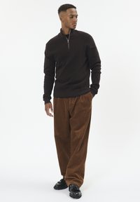 Man wearing brown corduroy pants, dark brown quarter-zip sweater, and black loafers standing against a white background.