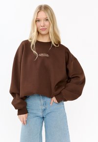 Blondynka pozująca z jedną ręką w kieszeni, ubrana w oversize'owy brązowy sweter z logo "MIRONS" oraz jasnoniebieskie jeansy z wysokim stanem.