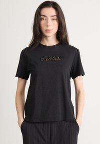 Čierne tričko s okrúhlym výstrihom vyrobené z bavlny, s krátkymi rukávmi, so zlatou vyšívanou logom "Calvin Klein" na hrudi.
