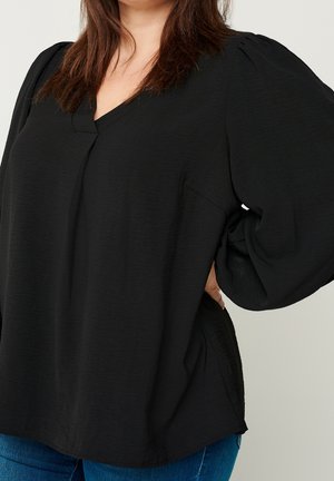 Femme portant un chemisier noir à manches longues avec un col en V et des manches bouffantes, associée à un jean bleu, debout, une main sur la hanche.