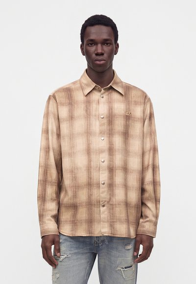 Homme portant une chemise à carreaux beige et marron à manches longues et un jean bleu clair déchiré, debout devant un fond blanc uni.