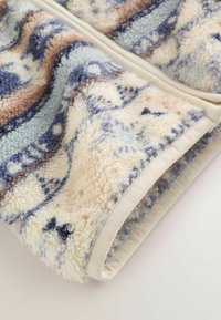 Nærbillede af et foldet blødt fleece tæppe med blå, beige og hvide geometriske mønstre og en beige stofkant.