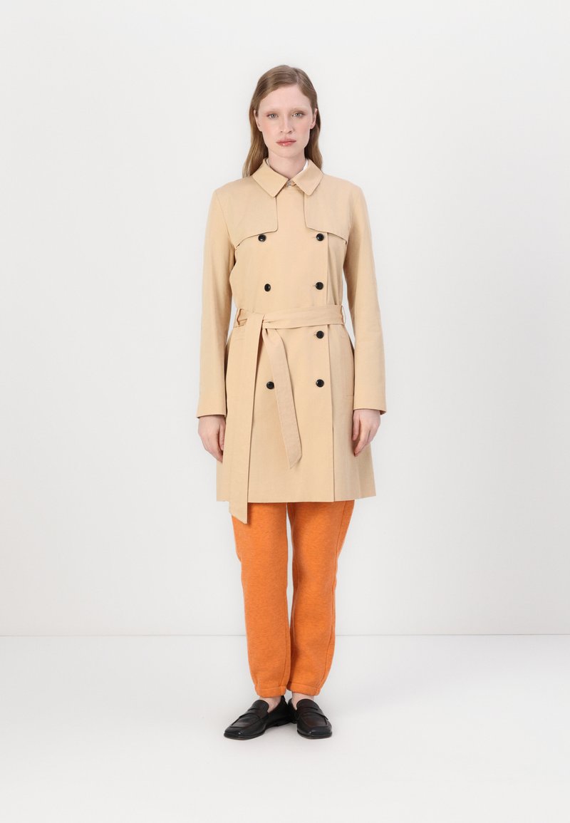 Manteau trench beige à double boutonnière avec des boutons noirs et une taille ceinturée, associé à un pantalon de survêtement orange et des chaussures noires à enfiler.