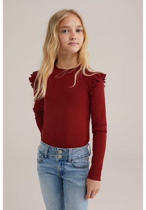 Young girl with long blonde hair wearing a burgundy long-sleeve top with ruffled shoulders and light blue jeans, standing against a plain background.
