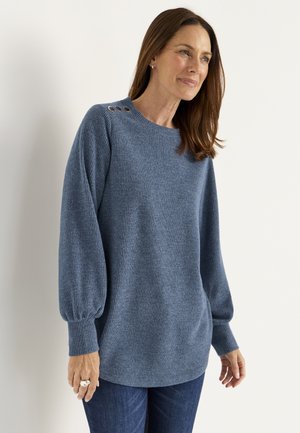Frau in einem blauen Strickpullover mit Knopfdetails an der Schulter und dunkelblauen Jeans, die vor einem schlichten weißen Hintergrund steht.