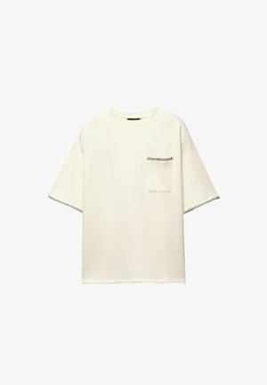 Bela oversize majica iz bombaža z kratkimi rokavi, s prsno žepom in kontrastnim črnim šivom ob robovih.