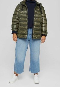 Giubbotto puffer verde oliva con finitura lucida, chiusura con zip e cappuccio. Abbinato a jeans a gamba larga blu chiaro e sneakers bianche.