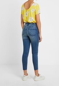 Top floral amarillo con mangas cortas, combinado con jeans ajustados azules de cintura alta y zapatillas blancas. La vista trasera resalta la tela y el ajuste.