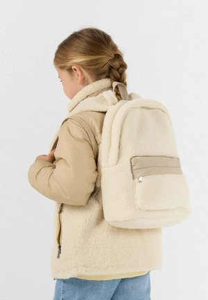 Niño con trenzas vestido con una chaqueta sherpa beige y una mochila sherpa a juego, mirando hacia la izquierda sobre un fondo liso.