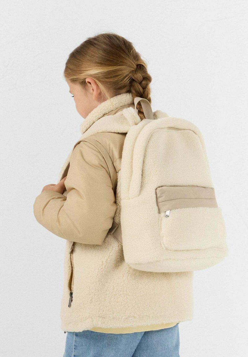 Kind met gevlochten haar, gekleed in een beige sherpa-jas en bijpassende sherpa-rugzak, kijkt naar links tegen een effen achtergrond.