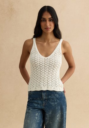 Giovane donna con capelli lunghi e scuri che indossa un top bianco senza maniche lavorato a maglia e jeans blu con motivo floreale, in piedi davanti a uno sfondo beige.