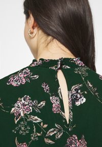Robe florale verte avec des motifs de fleurs roses et crème, dotée d'un col froncé et d'un détail en forme d'œillet dans le dos.