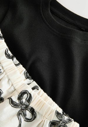 Melna ribota augšdaļa ar krēmkrāsas auduma apakšdaļu, uz kuras redzams melns lentu raksts. Augšdaļai ir apaļa kakla izgriezuma forma, bet apakšdaļa ir savākta.