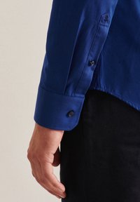 Detailní záběr na levou ruku a zápěstí osoby oblečené v modré košili s dlouhým rukávem a zapnutým manžetem, s černými kalhotami.