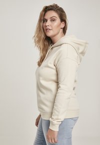 Krémová mikina s klokaním vreckom, nastaviteľnou kapucňou s šnúrkou a rebrovanými manžetami. Skombinovaná so svetlo modrými skinny džínsami.