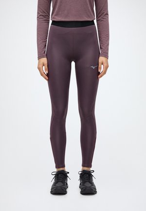 Le modèle se tient face à l’objectif, vêtu d’un legging de sport prune foncé, d’un haut à manches longues assorti et de baskets noires, sur un fond uni.