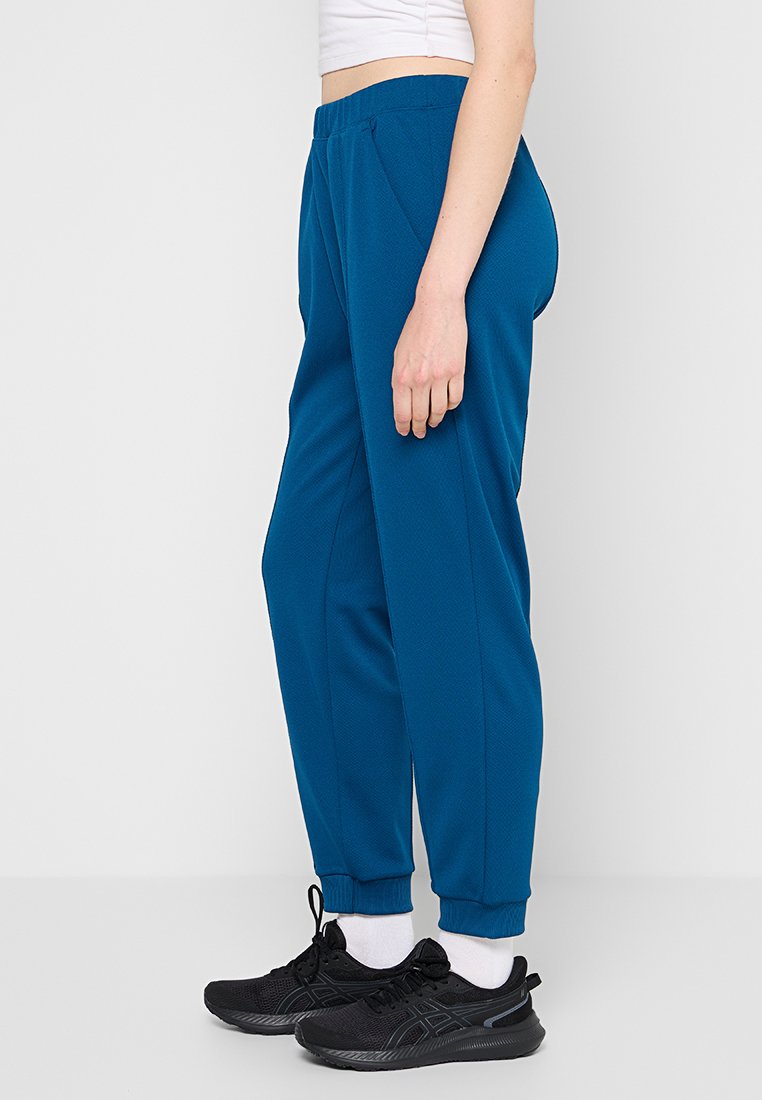Türkise Jogginghosen mit strukturiertem Finish, elastischem Bund, Seitentaschen und gerippten Bündchen, kombiniert mit schwarzen Sportschuhen.
