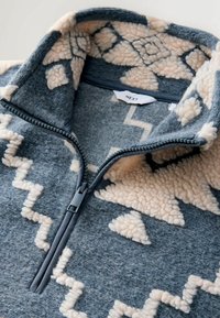 Fleece pullover with a zippered collar. Blue-gray fabric features cream geometric patterns. Soft texture and contrast zip detail.