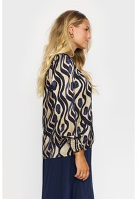 Beige blus med marinsjöl vågmönster. Har långa ärmar med samlade manschetter. Slät textur och avslappnad passform.