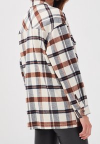 Chemise en flanelle à carreaux dans des tons crème, marron et marine. Elle possède un col pointu, des poignets à boutons et une coupe décontractée avec une poche plaquée.