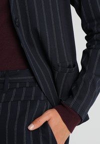 Marinblå kavaj med pinstripe-mönster och strukturerad yta, prydd med vit sticksöm, i kombination med en burgundy ribbad tröja. Sidofickdetalj synlig.