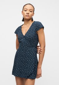 Γυναίκα με πλεγμένα μαλλιά που φοράει φόρεμα τύπου wrap με κοντά μανίκια σε ναυτικό μπλε χρώμα και μικρό λευκό ανθισμένο σχέδιο σε απλό φόντο.