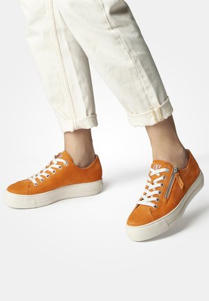 Sneakers in suede arancione con lacci bianchi, suole in gomma e dettagli con zip laterale. Pantaloni crema arrotolati mettono in risalto il design delle scarpe.