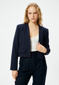 Laivastonsininen, lyhyt bleiseri, jossa on pystysuorat valkoiset raidat, yksittäinen etutasku ja avoin etuosasuunnittelu. Kangas on sileä ja siinä on hieman kiiltoa.