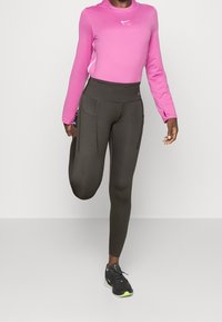 Camiseta deportiva rosa de manga larga con escote redondo, combinada con leggings oscuros de tiro alto y verde oscuro que tienen bolsillos laterales. Zapatillas negras con acentos verdes.