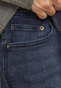 Dark blue denim jeans with a subtle texture, featuring metal rivets on pockets and contrasting yellow stitching along the seam.