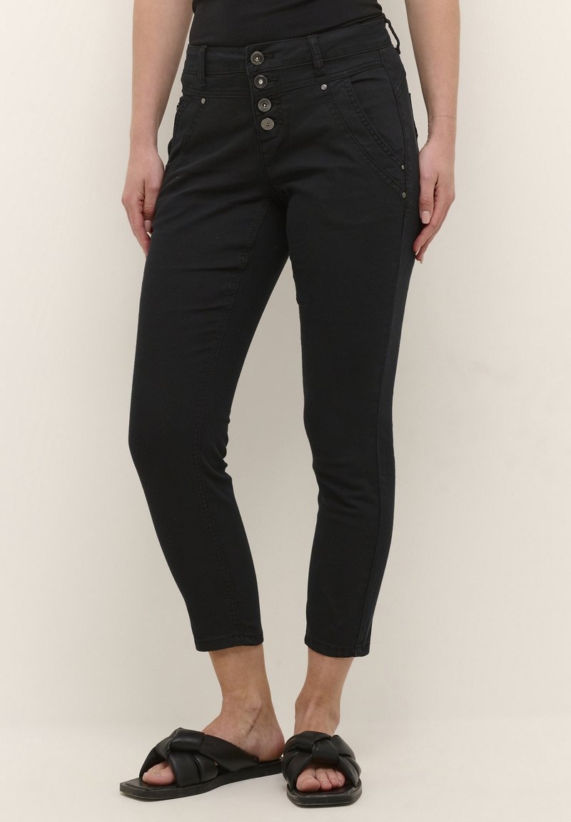 Pantalones negros de corte ajustado con cierre de cinco botones, bolsillos inclinados y costuras decorativas. La tela tiene una textura suave con un ligero estiramiento.