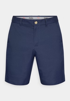 Pantalones cortos azul marino con textura suave, cuentan con un botón dorado, trabillas para cinturón y un logo de Puma en la cintura. Diseño clásico y longitud hasta la mitad del muslo.