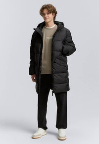 Schwarzer Steppmantel mit langer Silhouette, Kapuze und Frontreißverschluss. Verfügt über Seitentaschen und eine glatte, gepolsterte Textur. Getragen über einem beigen Sweatshirt.