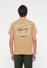 Mees seisab kaamera poole seljaga, seljas beež T-särk, millel on "Tommy Jeans NYC 1985" logo ja tumerohelised püksid, valge taustaga.