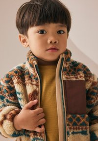 Jeune enfant avec des cheveux courts et sombres, portant une veste en polaire à motifs et un t-shirt jaune moutarde, tenant la fermeture éclair de la veste.