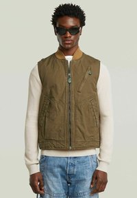 Giubbotto senza maniche verde oliva con zip frontale, due tasche laterali e colletto marrone. Indossato sopra un maglione color crema.