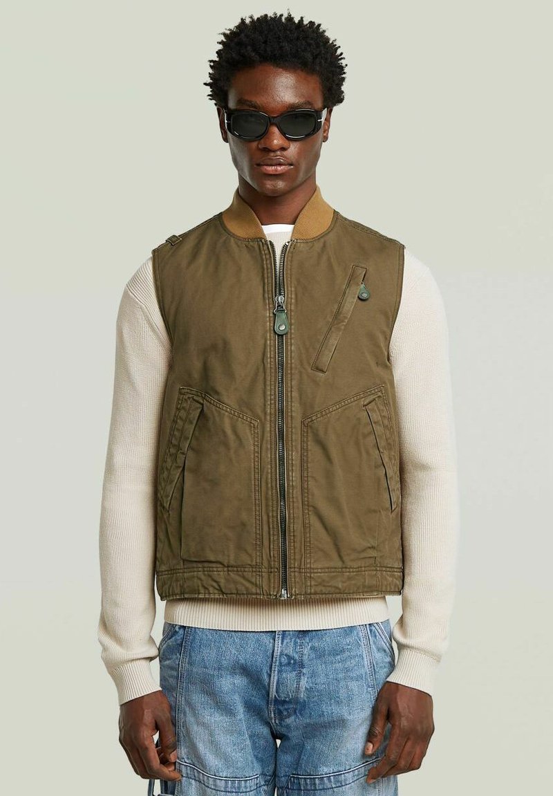 Giubbotto senza maniche verde oliva con zip frontale, due tasche laterali e colletto marrone. Indossato sopra un maglione color crema.