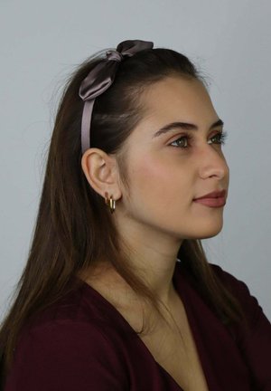 Fascia per capelli in raso marrone con fiocco, indossata da una persona con lunghi capelli castani, con orecchini a cerchio dorati e una maglia color bordeaux scuro.