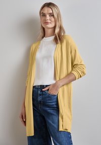 Gul stickad cardigan med öppen front, långa ärmar och sidsprund, kombinerad med en vit topp och blå jeans, mot en neutral bakgrund.