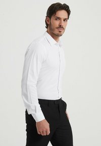 Chemise blanche à manches longues avec une coupe ajustée, devant boutonné et poignets doubles. Portée avec un pantalon noir, texture lisse et coupe moderne.